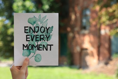 Woman holding card with phrase Enjoy Every Moment outdoors, closeup. Space for text