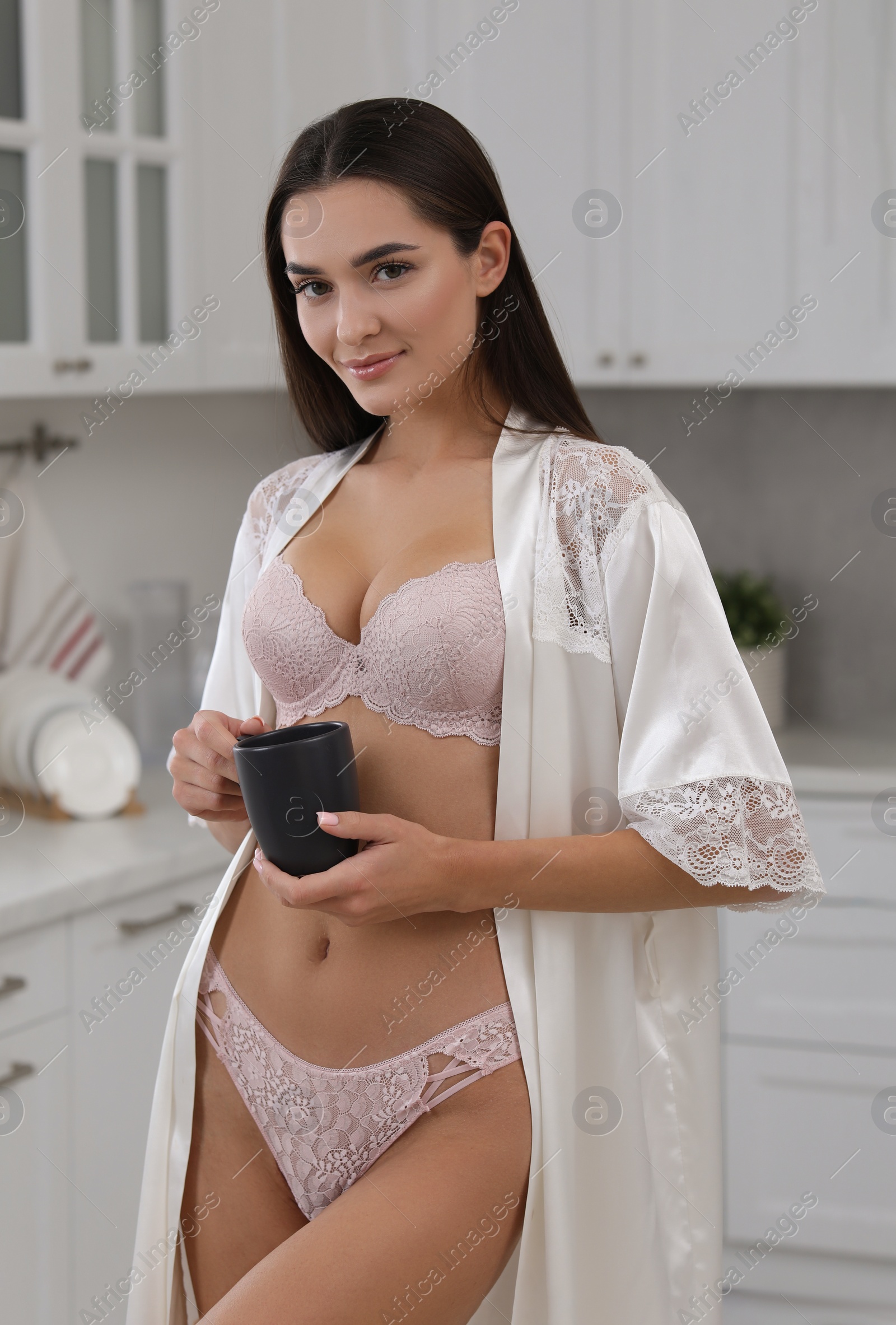 Photo of Young woman with cup wearing elegant underwear and robe in kitchen