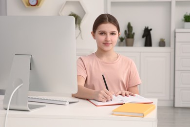 Photo of Cute girl writing in notepad while using computer at desk in room. Home workplace