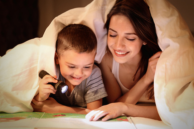 Photo of Mother and son with flashlight reading book under blanket at home