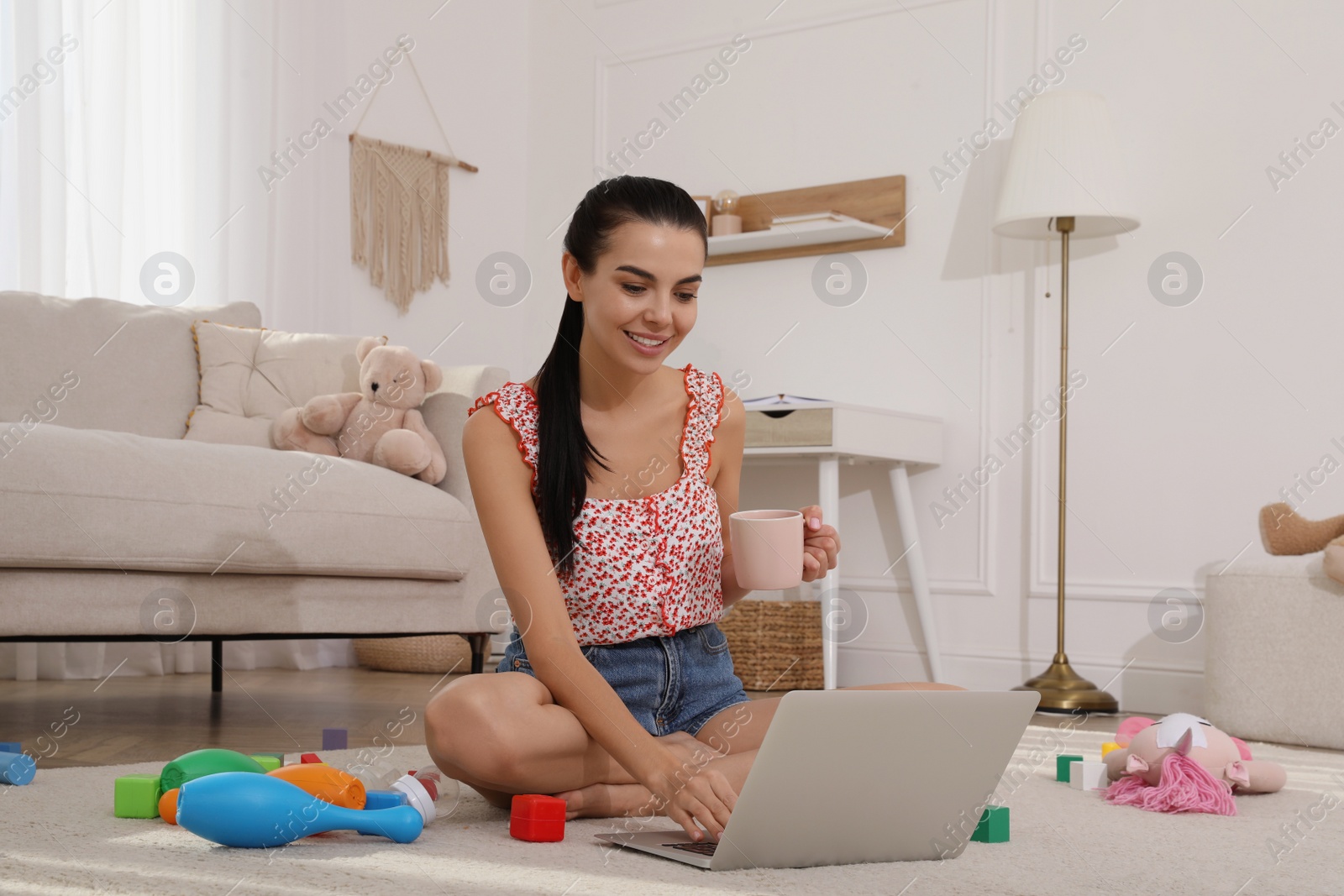 Photo of Happy young mother with cup of coffee and laptop resting in messy living room