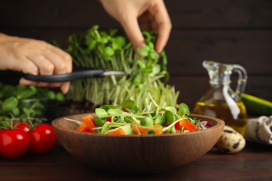 Photo of Woman cutting fresh organic microgreen at wooden table, focus on bowl of delicious salad