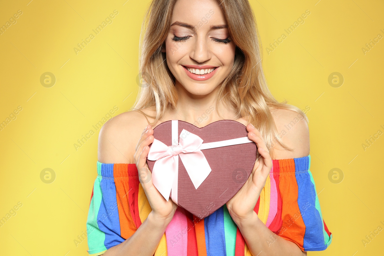 Photo of Portrait of beautiful smiling girl with heart shaped gift box on yellow background. International Women's Day