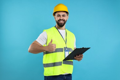 Photo of Engineer in hard hat holding clipboard and showing thumb up on light blue background