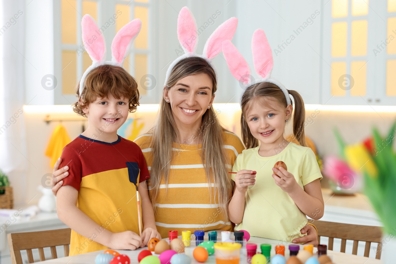 Photo of Painting Easter eggs. Happy mother and her cute children with bunny ears at table in kitchen