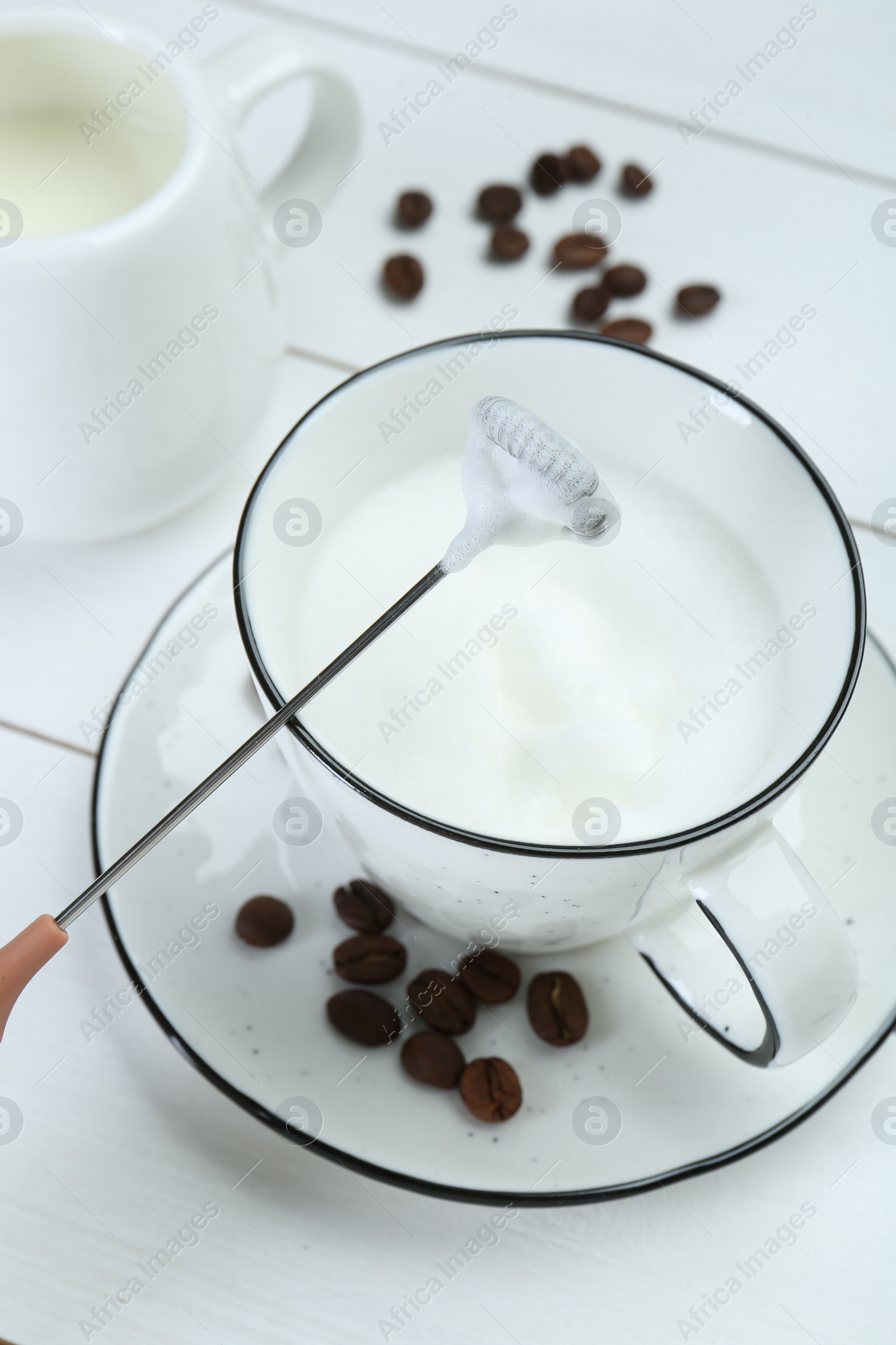 Photo of Mini mixer (milk frother), whipped milk and coffee beans on white wooden table, closeup