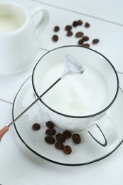 Mini mixer (milk frother), whipped milk and coffee beans on white wooden table, closeup