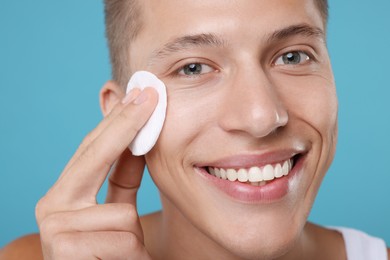 Handsome man cleaning face with cotton pad on light blue background, closeup