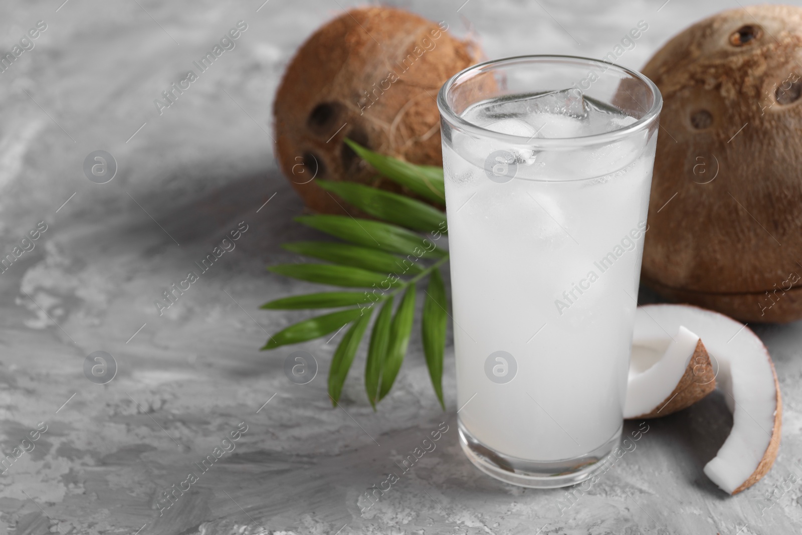 Photo of Glass of coconut water with ice cubes, palm leaf and nuts on grey table. Space for text