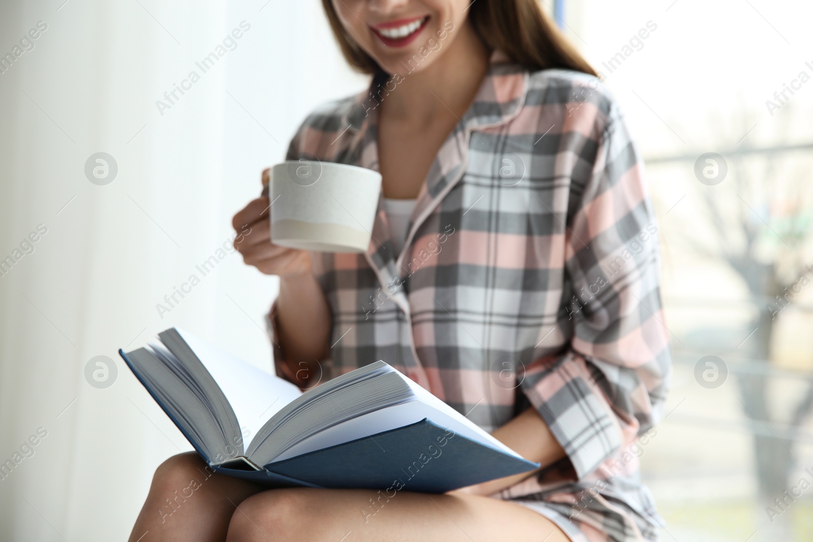 Photo of Young woman with cup of coffee reading book near window at home, closeup