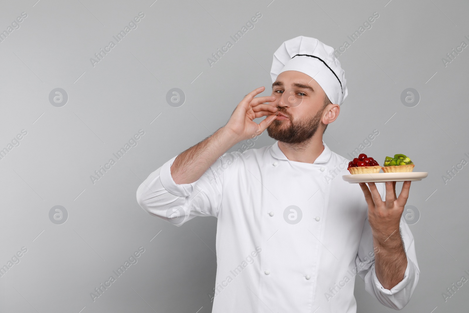 Photo of Happy professional confectioner in uniform with tartlets showing delicious gesture on light grey background. Space for text