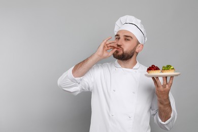 Happy professional confectioner in uniform with tartlets showing delicious gesture on light grey background. Space for text