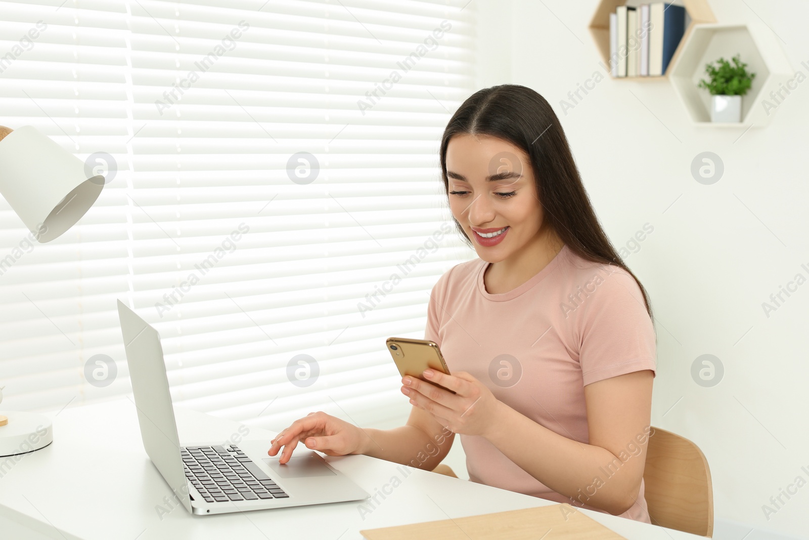 Photo of Home workplace. Happy woman with smartphone near laptop at white desk in room