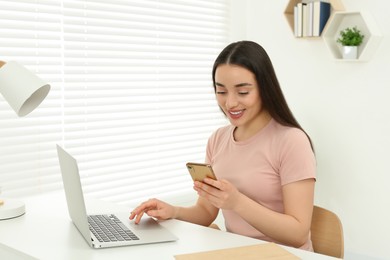 Photo of Home workplace. Happy woman with smartphone near laptop at white desk in room