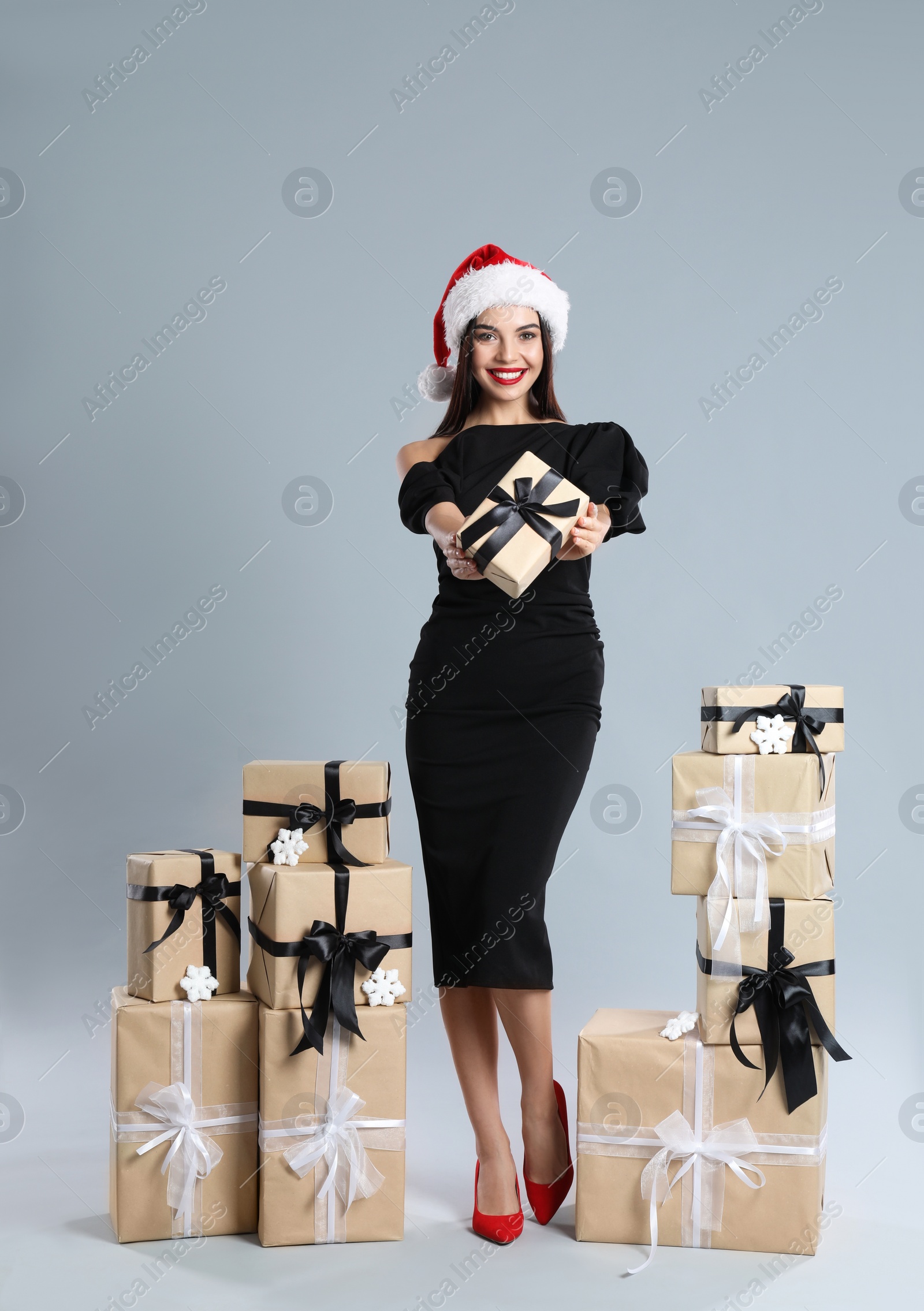 Photo of Woman in black dress and Santa hat with Christmas gifts on grey background