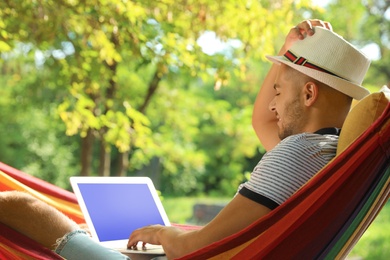 Young man with laptop resting in comfortable hammock at green garden