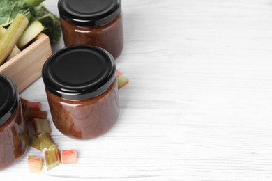 Photo of Jars of tasty rhubarb jam and cut stalks on white wooden table, space for text