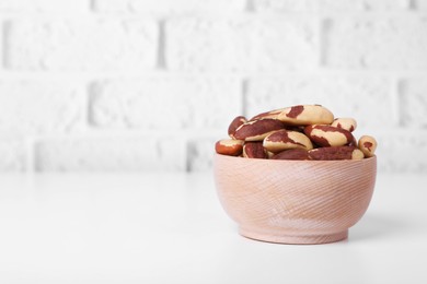 Photo of Bowl with tasty Brazil nuts on white table against brick wall. Space for text