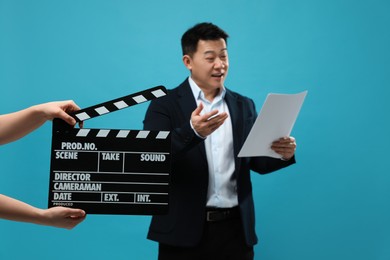 Photo of Happy asian actor performing role while second assistant camera holding clapperboard on light blue background, selective focus. Film industry