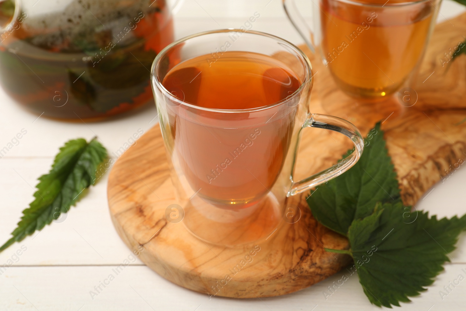 Photo of Glass cup of aromatic nettle tea and green leaves on white wooden table, closeup