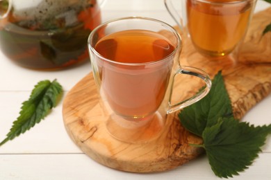 Photo of Glass cup of aromatic nettle tea and green leaves on white wooden table, closeup