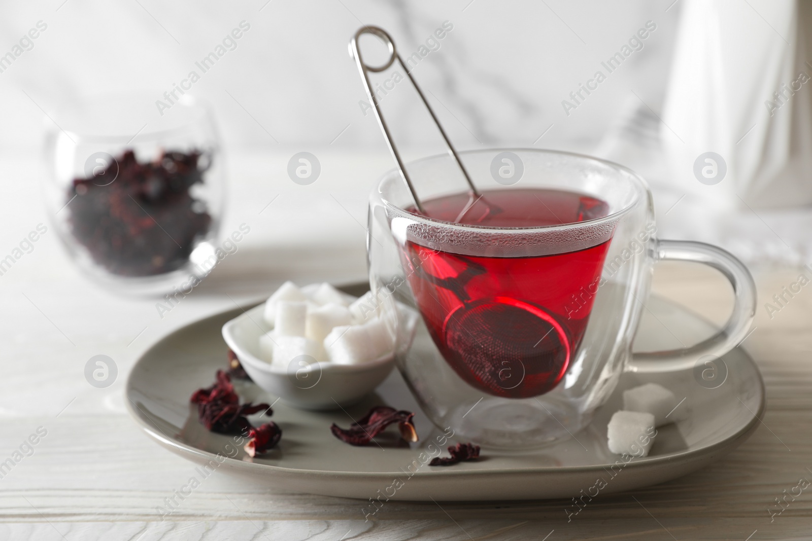 Photo of Delicious hibiscus tea in cup, sugar cubes and dry roselle petals on white wooden table, closeup