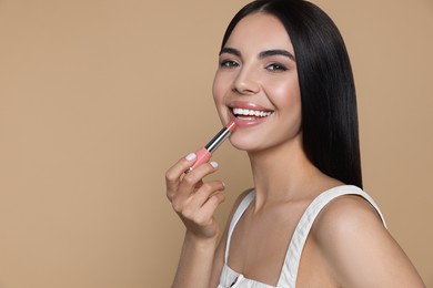 Beautiful young woman applying nude lipstick on beige background, space for text