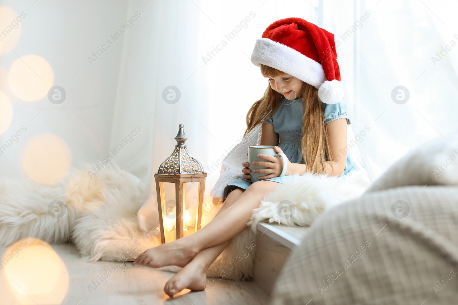 Photo of Cute little child in Santa hat with cup of cocoa sitting on windowsill at home. Christmas celebration