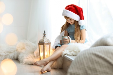 Photo of Cute little child in Santa hat with cup of cocoa sitting on windowsill at home. Christmas celebration