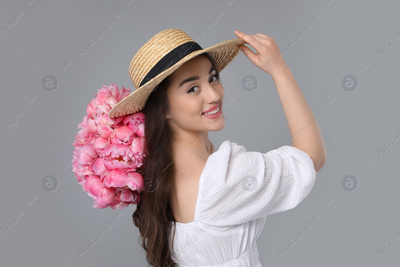 Photo of Beautiful young woman in straw hat with bouquet of pink peonies against grey background