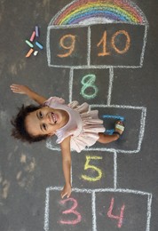 Little African American girl and colorful hopscotch drawn with chalk on asphalt outdoors, top view. Happy childhood