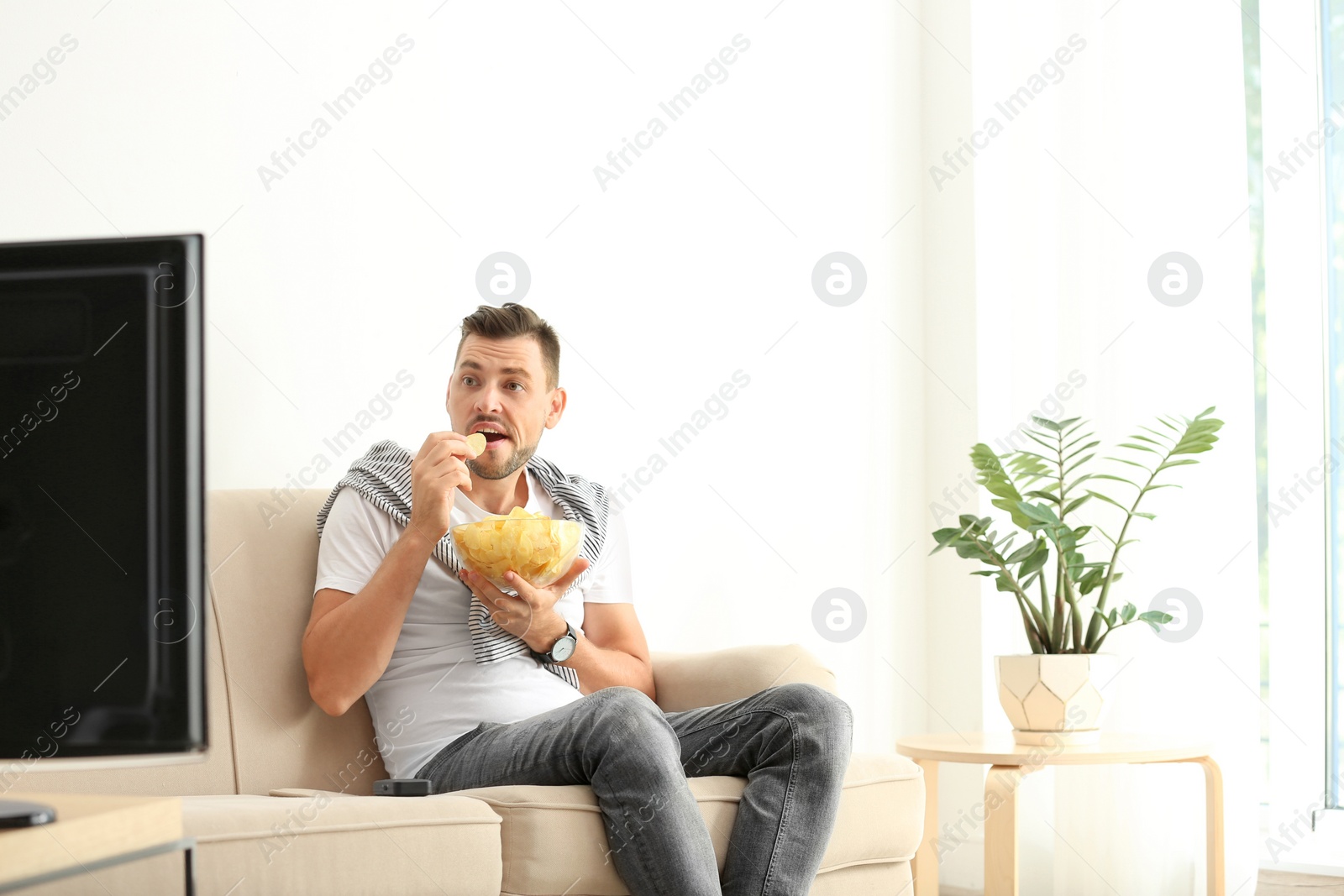 Photo of Man with bowl of potato chips watching TV on sofa in living room