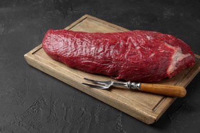 Photo of Wooden board with piece of raw meat and fork on black table