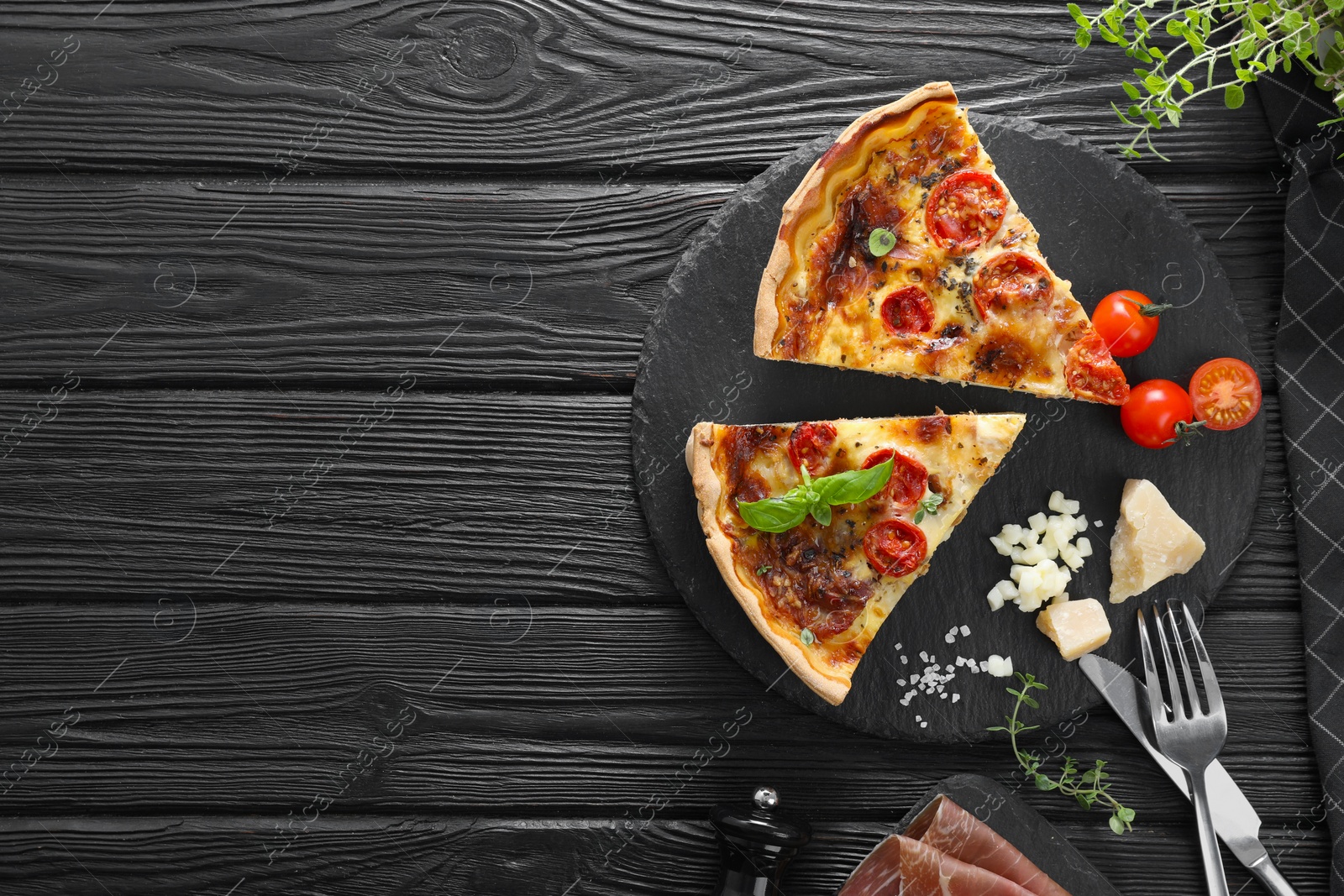 Photo of Pieces of delicious homemade quiche with prosciutto and ingredients served on black wooden table, flat lay. Space for text