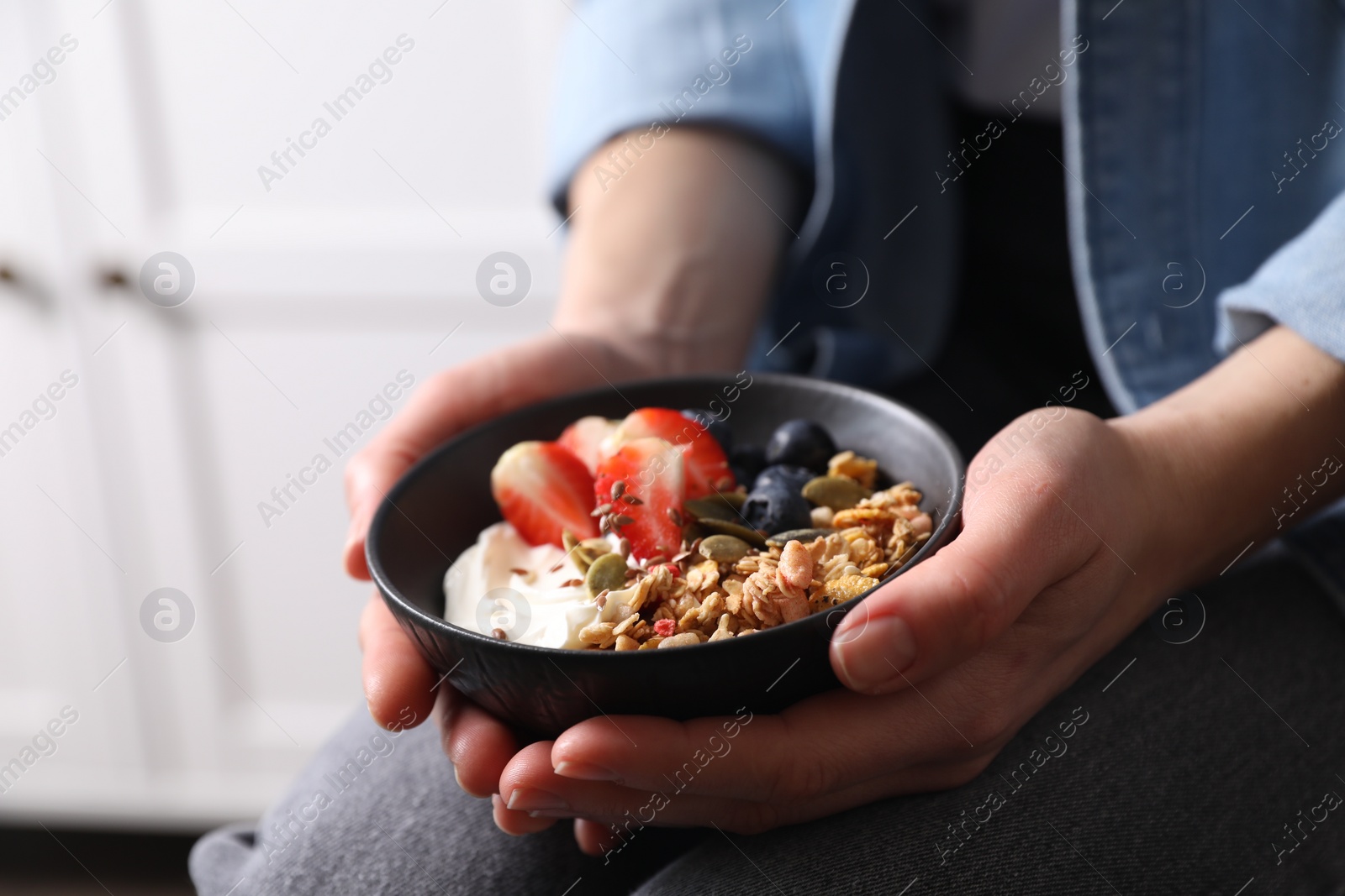 Photo of Woman holding bowl of tasty granola with berries, yogurt and seeds indoors, closeup