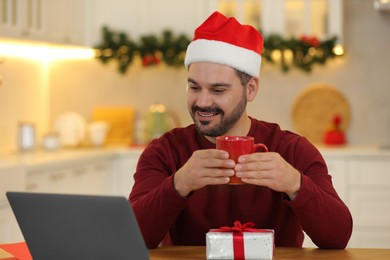 Celebrating Christmas online with exchanged by mail presents. Happy man with cup of drink and gift box during video call on laptop at home