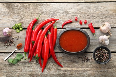 Photo of Spicy chili sauce in bowl and ingredients on wooden table, flat lay