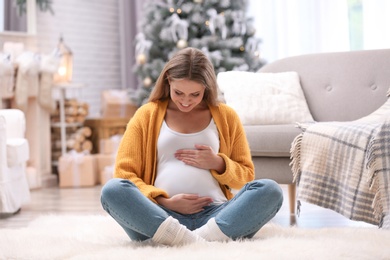 Photo of Young pregnant woman sitting on floor in room decorated for Christmas