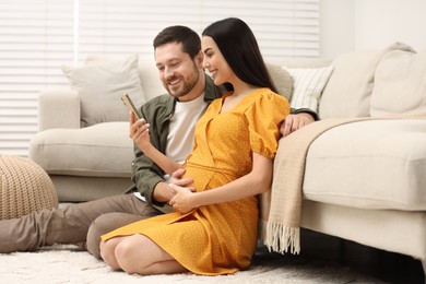 Photo of Happy pregnant woman with her husband using smartphone on soft carpet at home