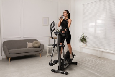 Photo of Happy young woman drinking water during training on elliptical machine at home