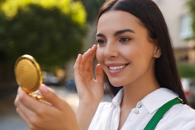 Beautiful young woman looking at herself in cosmetic pocket mirror outdoors