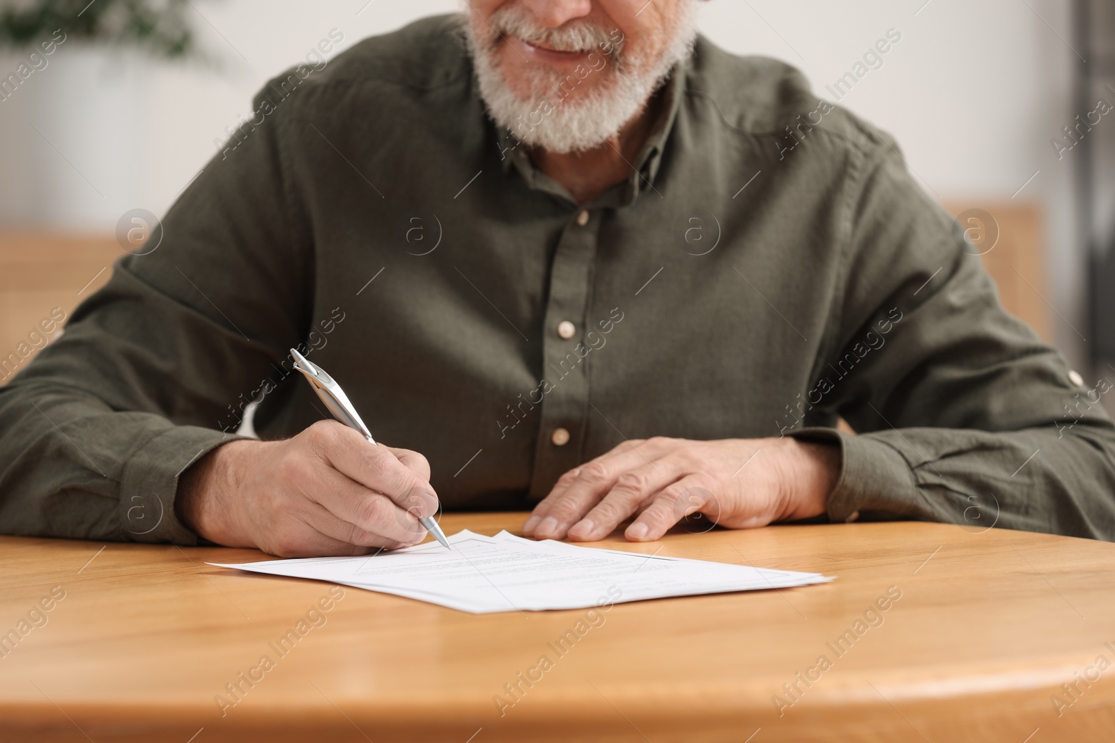 Photo of Senior man signing Last Will and Testament at wooden table indoors, closeup