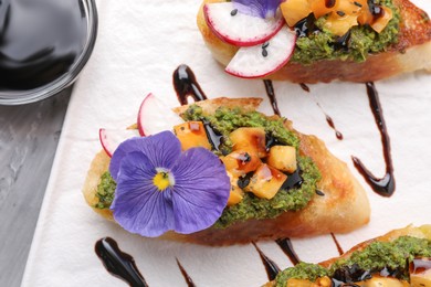 Photo of Delicious bruschettas with pesto sauce, tomatoes, balsamic vinegar and violet flower on table, above view