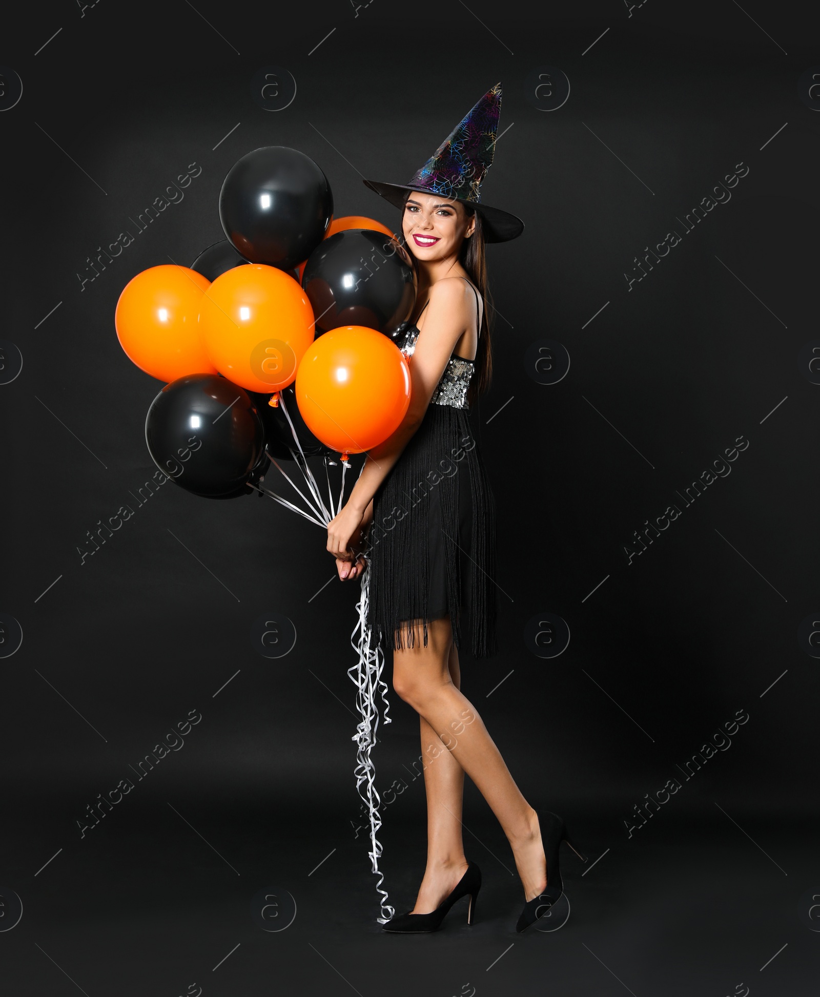 Photo of Beautiful woman wearing witch costume with balloons for Halloween party on black background