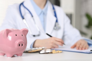 Doctor making notes at white table indoors, focus on piggy bank