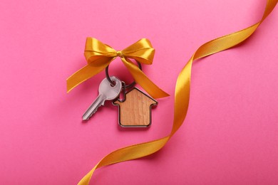 Key with trinket in shape of house, bow and ribbon on pink background, flat lay. Housewarming party