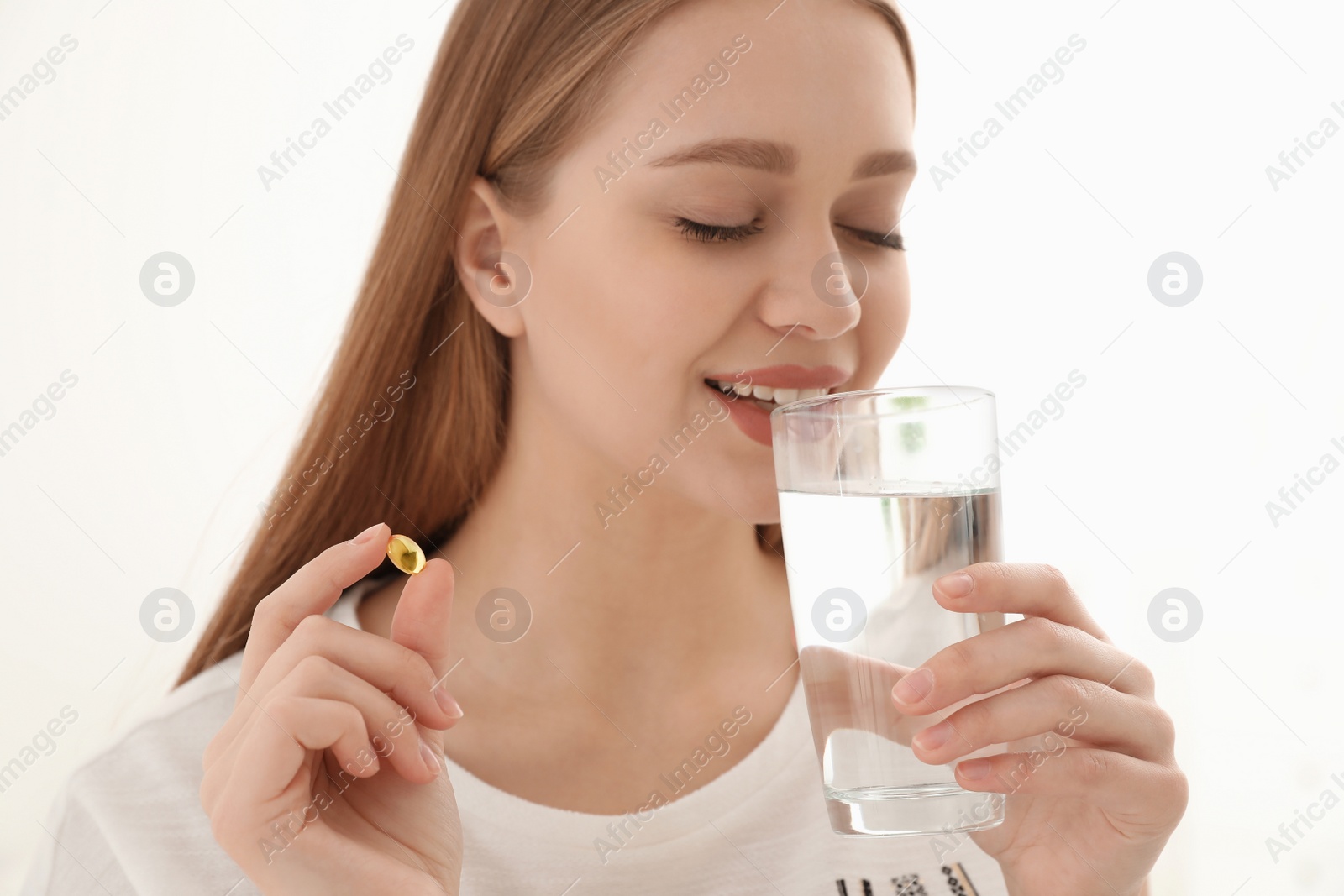 Photo of Young woman with glass of water taking vitamin capsule on light background