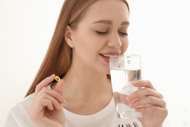 Young woman with glass of water taking vitamin capsule on light background