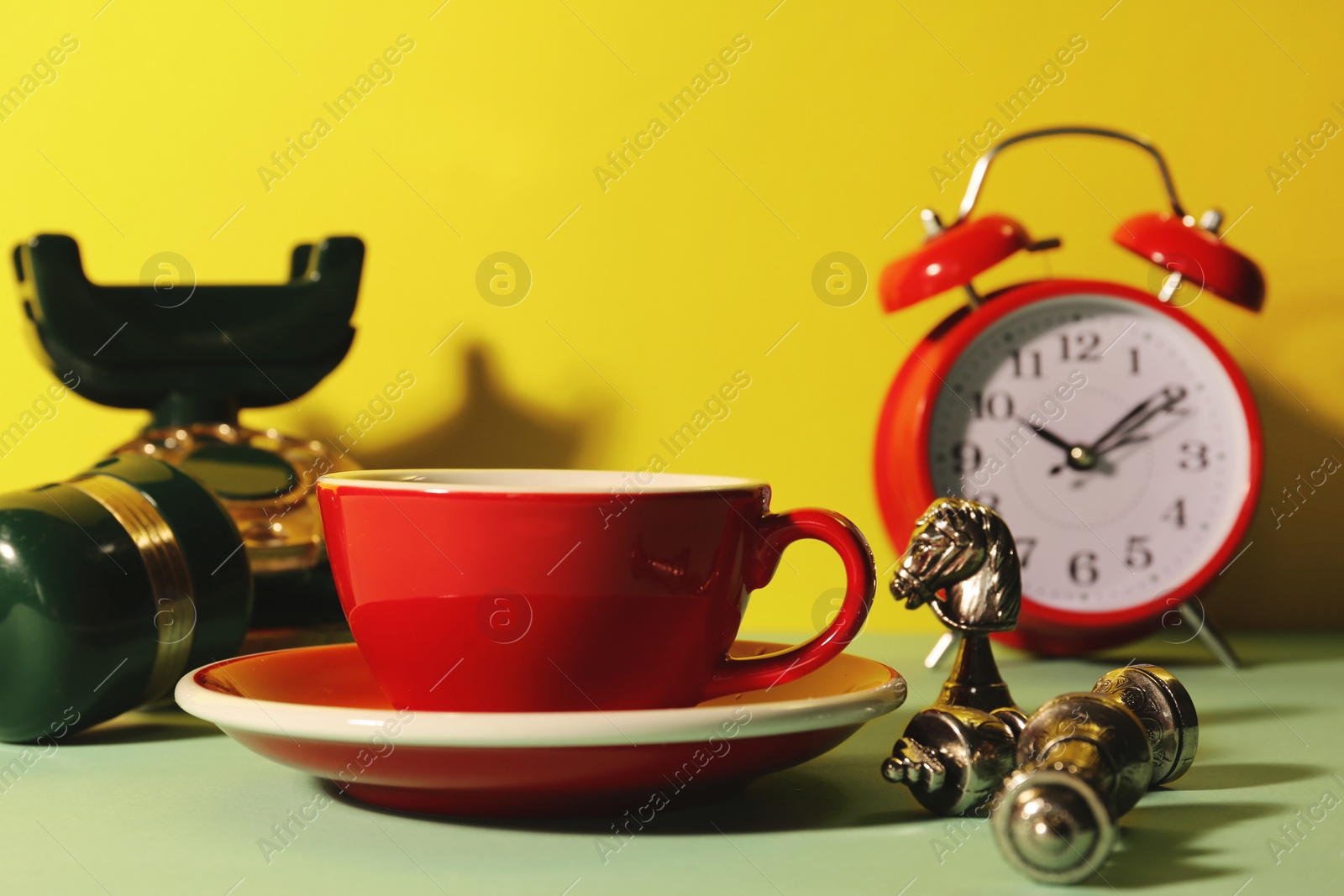 Photo of Red cup with saucer, alarm clock, vintage telephone and chessmen on light green table against yellow background
