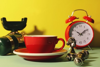 Red cup with saucer, alarm clock, vintage telephone and chessmen on light green table against yellow background